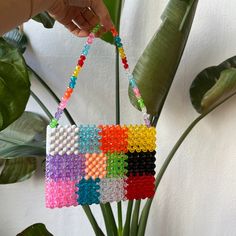 a hand holding a multicolored beaded purse hanging from a palm tree in front of a white wall