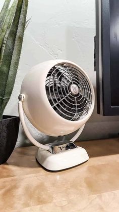 a white fan sitting on top of a wooden table next to a computer monitor and cactus