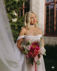 a woman in a white wedding dress holding a pink and red bouquet standing next to a building