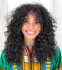 a woman with long curly hair wearing a green shirt and smiling at the camera while standing in front of a white door