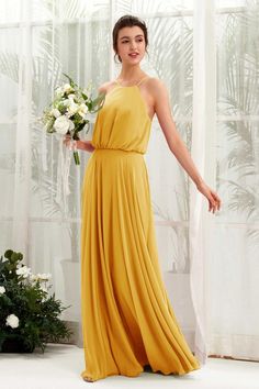 a woman in a long yellow dress standing next to a white flower arrangement and holding a bouquet