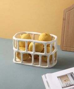 three lemons in a white basket sitting on a table next to an open magazine