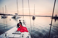 a man and woman sitting on the bow of a sailboat at sunset with boats in the background