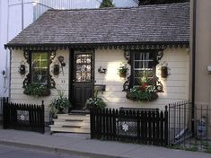 a small white house with potted plants on the front and side windows, along with a black fence