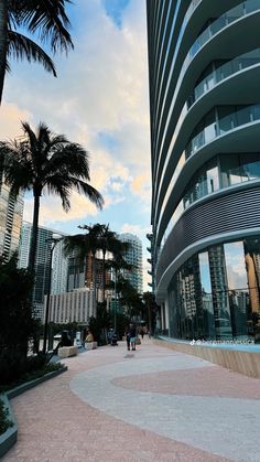 people are walking in front of a building with palm trees on the sidewalk and buildings behind it