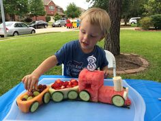 a young boy sitting at a table with a train made out of watermelon and cucumbers