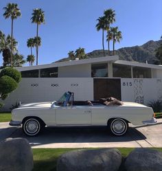 an old white convertible car parked in front of a house with palm trees and rocks