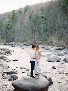 an engaged couple standing on rocks in the middle of a river