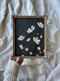 a person holding up a framed painting with white flowers on black and gold foiled paper