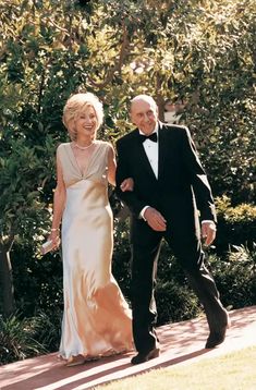 an older man and woman in formal wear walking down a path together, smiling at the camera