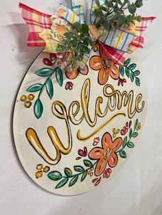 a welcome sign hanging on the side of a wall with flowers and ribbon around it