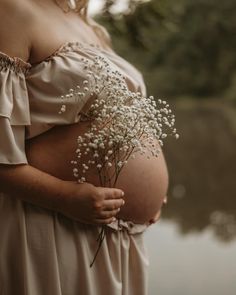 a pregnant woman holding flowers in her belly while standing next to a body of water