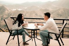 a man and woman sitting at a table on top of a mountain with mountains in the background