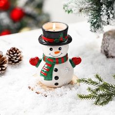 a snowman candle holder sitting in the snow next to pine cones and christmas decorations