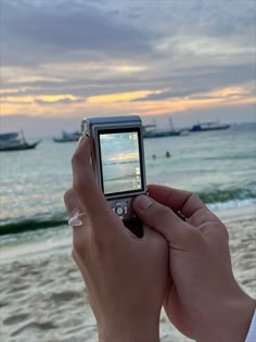 a person holding a cell phone up to their face on the beach with boats in the water