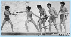 four men in bathing suits standing on the edge of a boat