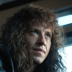 a man with long curly hair and blue eyes looks off into the distance while sitting in a car