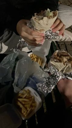 two people holding sandwiches and french fries in their hands while sitting on the ground outside