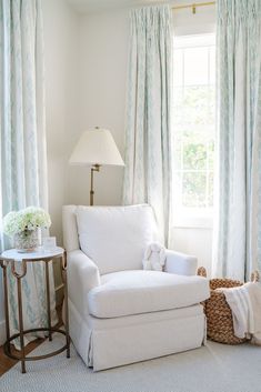 a white chair sitting in front of a window next to a table with a lamp on it