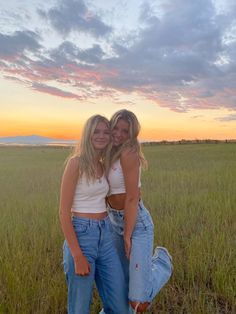 two young women standing next to each other in a field at sunset with the sun setting behind them