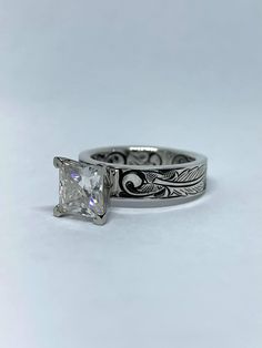 a white gold ring with a princess cut diamond in the center and an intricate filigree design on the band