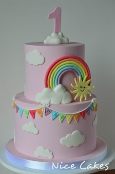 a pink birthday cake decorated with clouds, rainbows and the number one on top