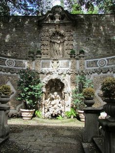 an outdoor fountain surrounded by stone pillars and potted plants in front of a brick wall