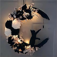 a black cat sitting in front of a wreath with white flowers and lights on it