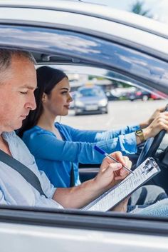 a man and woman sitting in the drivers seat of a car, writing on a clipboard