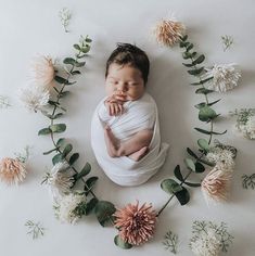 a newborn baby wrapped in a white wrap surrounded by flowers and greenery on a white background