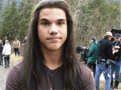 a young man with long hair standing in front of a group of people on the side of a road