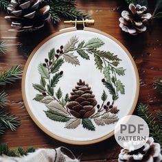 an embroidered christmas wreath with pine cones and evergreen needles is displayed on a wooden table