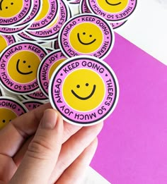 someone holding up some stickers with smiley faces on them in front of a pink and purple background