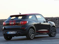 the rear end of a small black car parked in front of a stone wall and fence