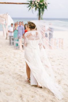 the bride is walking down the beach with her veil blowing in the wind as people watch