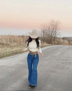 a woman walking down the middle of a road wearing a cowboy hat and blue jeans