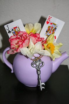 a purple teapot filled with flowers and playing cards on top of a black table