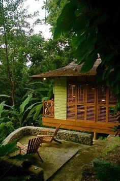 a small wooden cabin in the middle of some trees and greenery with two chairs