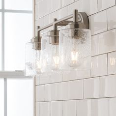 a bathroom light with three lights on the wall and white tile behind it, in front of a window