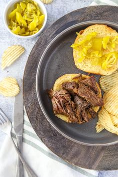 a steak sandwich with potato chips on a plate next to a bowl of pickles