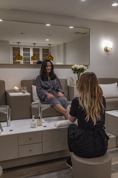 a woman sitting on a stool in front of a mirror next to another woman with long hair