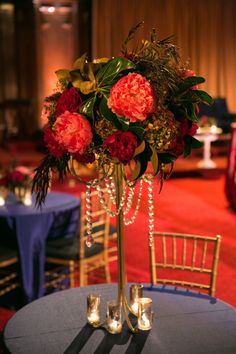 a tall vase with red flowers and greenery on top of a blue table cloth