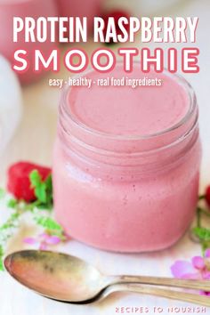 Photo of a small glass jar filled with a pink smoothie, sitting next to a fresh raspberries, pink flowers and white and green flowers around it, silver spoons in front of it an a couple small jars filled with the pink smoothie in the background with text that says Easy Protein Raspberry Smoothie.