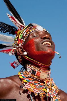 a man with red paint on his face and feathers around his neck, standing in front of a blue sky