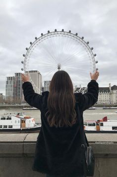 a woman standing in front of the london eye looking out over the river thames with her hands up