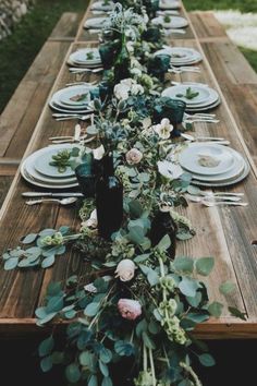 a long wooden table with plates and flowers on it is set for an outdoor dinner