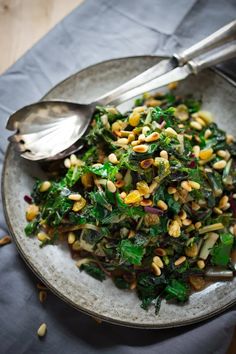 a white plate topped with greens and other vegetables next to silver spoons on top of a table