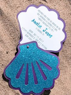 a blue and purple scallop shell on the sand with a wedding card attached to it