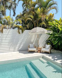 two chairs under an umbrella next to a swimming pool with palm trees in the background