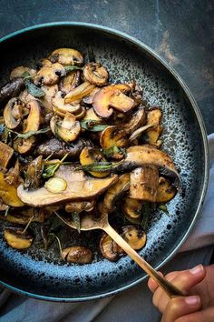 a pan filled with mushrooms and spinach on top of a blue table cloth next to a person holding a wooden spoon